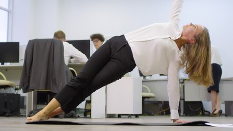 Wide shot of blonde barefoot businesswoman doing yoga on mat in office while her coworkers working at desks on background