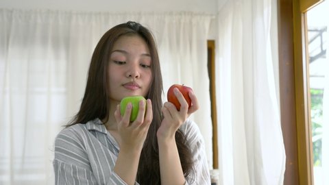 portrait of a young lovely pretty asian girl biting an green apple in white kitchen in morning. Her hands holding green and red apple that made her strong and healthy