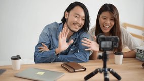 Slow motion, Young Asian man and woman are waving the hand with the camera which standing on the wood table. the couple man and woman sitting in the home and record video clip. blogger and vlog concept.