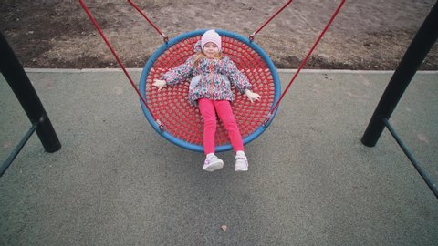 Little cute girl swinging on round swing with tight weave rope platform seat in park on cloudy day.