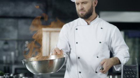 Chef cooking food with burning fire in slow motion. Focused chef throwing asian food in frying pan with flame at kitchen. Male chef preparing flambe dish with fire flames at restaurant