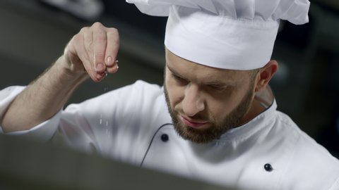 Portrait of chef salting food at kitchen. Focused chef cooking food in slow motion. Close up of bearded chef preparing meal at professional kitchen. Portrait of serious man working at restaurant