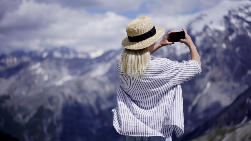 Young hipster girl in casual wear holding cellular phone in hands making photo of journey in mountains, female travel blogger taking picture of breathtaking nature landscape in vacations, slow motion