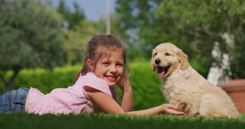 Slow motion of little girl lying on the lawn of a garden is cuddling and kissing a puppy of golden retriever dog.