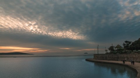 "Altocumulus" clouds moving time lapse during a sunset calm sea with island seen in a the far, silhouettes of people seen walking across the pedestrian sea bridge in the far. 