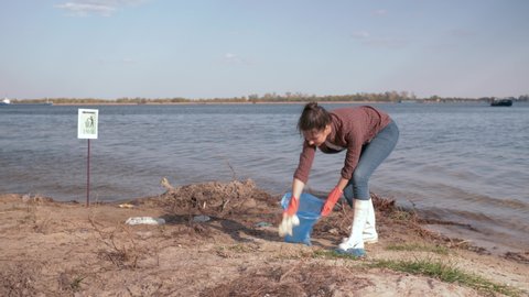 environmental solutions, happy girl volunteer collects plastic trash in garbage bag and gives positive gesture on polluted river beach near water