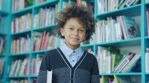 Zoom in of handsome mixed race little boy with curly hair smiling at camera standing near shelves in school library and holding book under his arm
