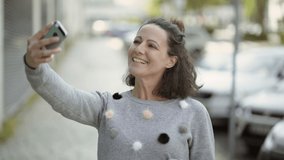 Happy middle aged woman having video call through phone. Smiling mature brunette holding smartphone in outstretched arm and talking with interlocutor. Communication and technology concept