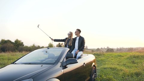 Handsome young couple in stylish glasses recording video, taking selfie photo using smart phone while sitting in the cabriolet on the nature. Dolly shot.