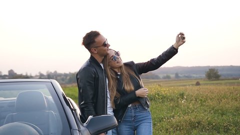 Handsome couple in love in black leather jacket taking selfie using smart phone standing near the car. cabriolet during sunset. Dolly shot.