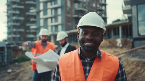Cheerful Afro-American builder looks straight to camera and smiles happily on working colleagues, unfinished construction background. Positive emotions, good mood. Male portrait