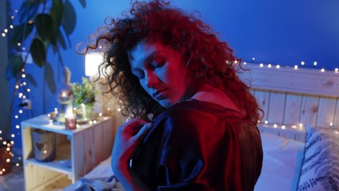 Pretty young Caucasian curly woman playfully covering her naked shoulders with black nightgown and seductively looks straight to camera in neon light. Sexy brunette lover in underwear lying on bed
