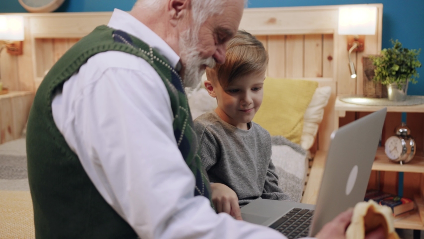 Glad smiling pretty boy in gray sweater looking at screen of smartphone in hands of granddaddy. Child watching funny videos. Cozy atmosphere in flat. Indoors.