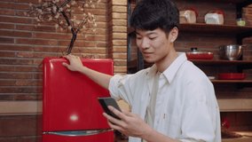 Cheerful young handsome chinese man making the video call and room tour via smartphone, being in the kitchen, opens fridge and laughs, joking. New generation technology, internet communication