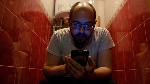 Sitting on the toilet, the man holds his smartphone to pass the time. Young man using mobile phone while sitting on the toilet at home.