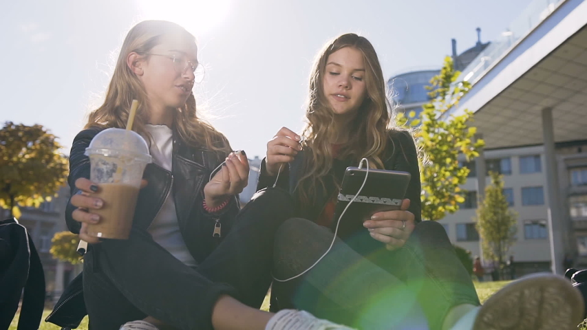 Sociable caucasian girlfriends using the tablet computer laughing at fun pictures on the tablet sitting outdoors in the park in sunny weather. Slow motion