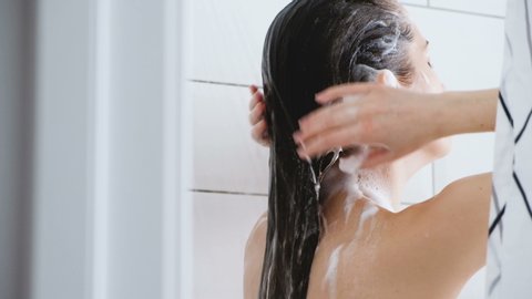 Young woman washes away the shampoo from her hair while standing under a warm shower stream. Concept of cleanliness of hair and body