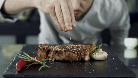 Chef salt grilled meat at stone cutting board. Closeup man hands salting steak in slow motion. Close up hands finishing ready steak. Closeup grill meat dish with vegetables at professional kitchen.