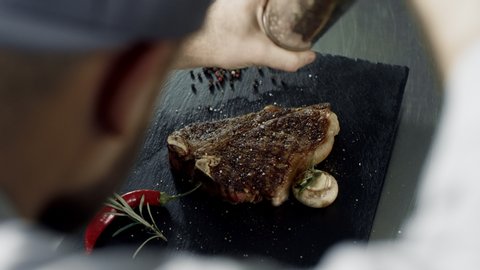 Chef peppering meat at stone cutting board. Closeup man hands peppering grilled streak in slow motion. Top view of chef hands decorating ready meat dish with pepper at professional kitchen