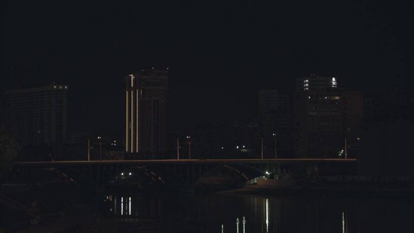 Night city landscape with a bridge above the river and moving car. Stock footage. Beautiful night city with embankment near the river on night sky background.