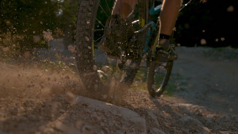 SLOW MOTION, LOW ANGLE, CLOSE UP, DOF: Bright sunbeams shine on a part of a gravel path as the downhill cyclist rides past camera and past the forest. Man brakes while riding e-bike down empty trail.