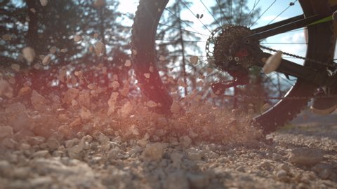 SLOW MOTION, SUN FLARE, CLOSE UP, LOW ANGLE: Cinematic shot of rocks flying as cyclist rides along a gravel path. Unrecognizable extreme bike rider brakes while mountain biking sending rocks flying.