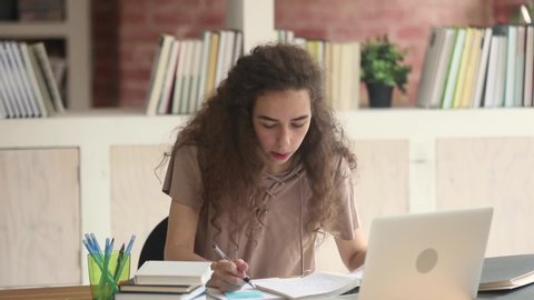 Focused teen college university student studying writing notes, serious high school girl prepare for test exam writing essay project coursework doing research homework learning in library with laptop