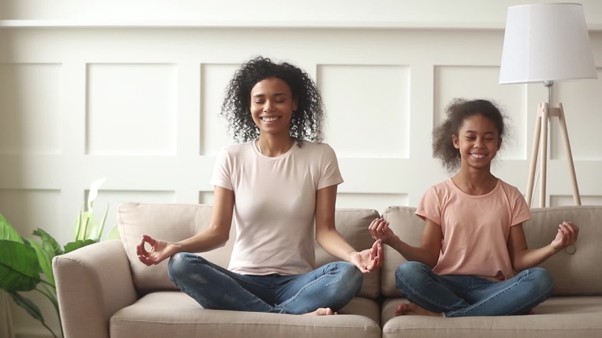 Happy family mindful african mom teaching cute funny kid daughter doing yoga exercise at home, calm healthy mixed race mother and little girl sitting in lotus pose on sofa meditating together | Shutterstock HD Video #1029713960