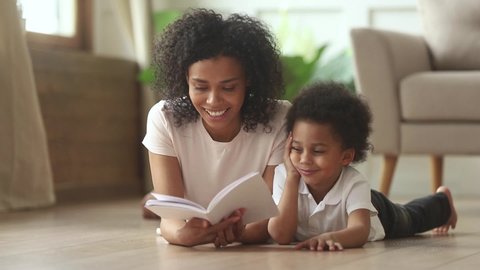 Loving african american mother telling fairy tale story to smart cute kid son laying on warm floor together, caring mixed race mom babysitter holding reading book to little child boy learning at home