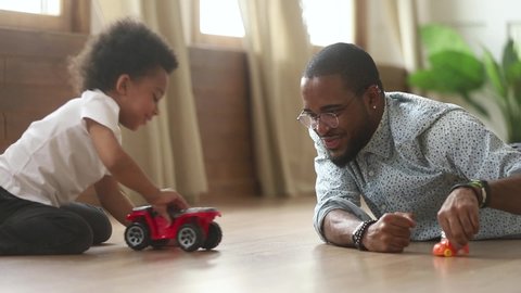 Happy african american cute little son playing toy cars with loving young dad babysitter, funny small kid son and black father having fun racing on warm floor at home, family daddy child men games