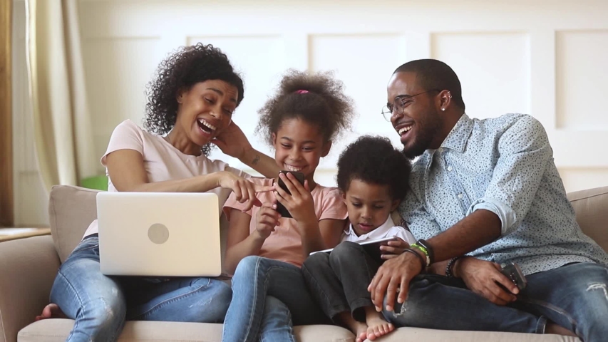 Happy african family parents and little children enjoy using devices together sit on sofa, technology addicted couple with kids having fun with laptop tablet phone at home, people gadget addiction Royalty-Free Stock Footage #1029713999