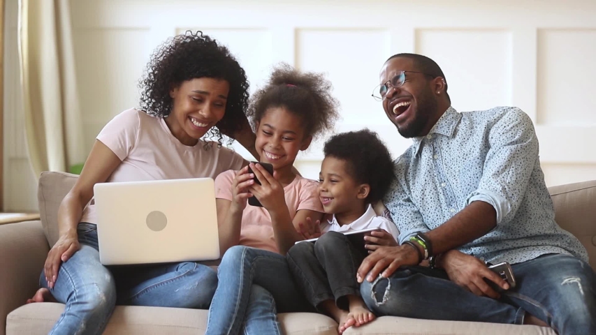 Happy african family parents and little children enjoy using devices together sit on sofa, technology addicted couple with kids having fun with laptop tablet phone at home, people gadget addiction | Shutterstock HD Video #1029713999