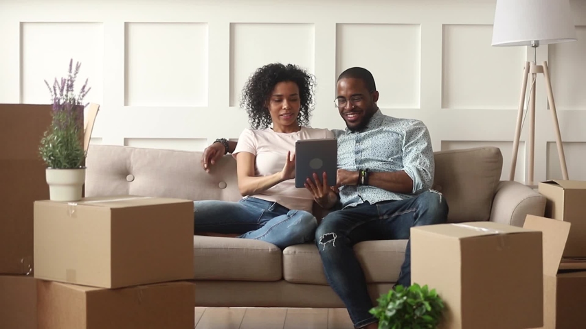 Happy african couple renters owners tenants sit on sofa use digital tablet on moving day in new house, black man and woman relax on couch with boxes discuss interior design online idea renovate home Royalty-Free Stock Footage #1029714002