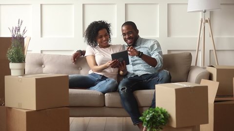 Happy african couple renters owners tenants sit on sofa use digital tablet on moving day in new house, black man and woman relax on couch with boxes discuss interior design online idea renovate home