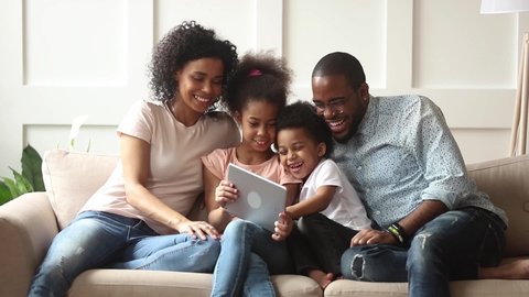 Cute little african children play online game using digital tablet having fun with parental control sit on sofa, happy black family laugh look at computer screen together enjoy do internet shopping