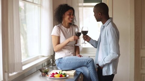 Happy young romantic african couple in love clinking glasses having fun talk laugh bonding in kitchen, affectionate black man and woman enjoy drink red wine celebrate anniversary together at home 