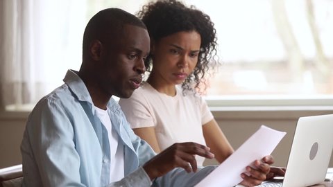 Serious african american couple talking doing paperwork using laptop together, focused family calculate pay bill rental payment online on computer holding paper at home office, mentor teaching intern