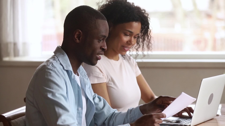 Happy african american young couple paying bank loan bills online at home, millennial black husband holding papers doing paperwork payment with wife using laptop planning budget sit at desk at home Royalty-Free Stock Footage #1029714080