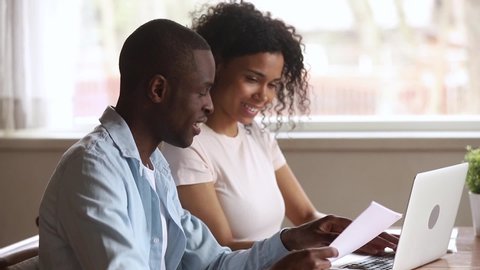 Happy african american young couple paying bank loan bills online at home, millennial black husband holding papers doing paperwork payment with wife using laptop planning budget sit at desk at home