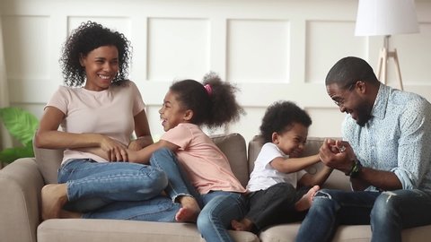 Happy african american family with kids laugh tickle sit on sofa, cheerful black parents and children having fun play at home, mom dad enjoy funny lifestyle activity with little cute son daughter