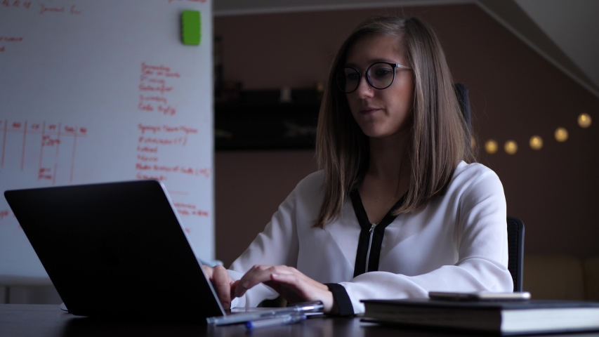 Young woman sits in home office with whiteboard behing the desk, works on laptop, types and scroll with concentration