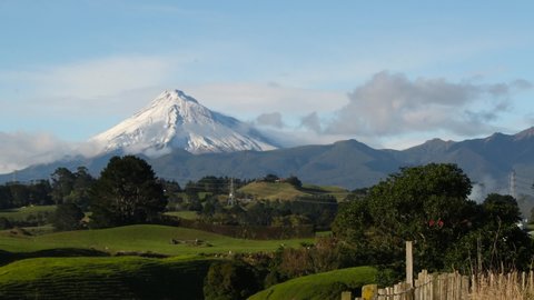 Snow capped mountain towers above green pasture in Taranaki, New Zealand on a sunny morning, TIME LAPSE, ZOOM.