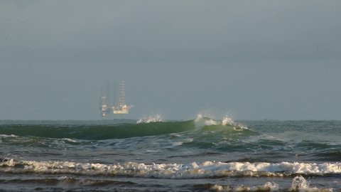 Waves crash on shore in front of oil rig in the distant ocean. STATIC
