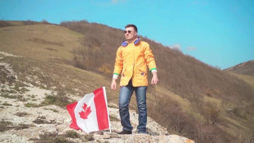 A man in a yellow jacket, blue jeans and glasses stands on a mountain and shows victory, the flag of Canada is set in the ground. Background of mountains and sky. Flag of Canada develops in the wind.