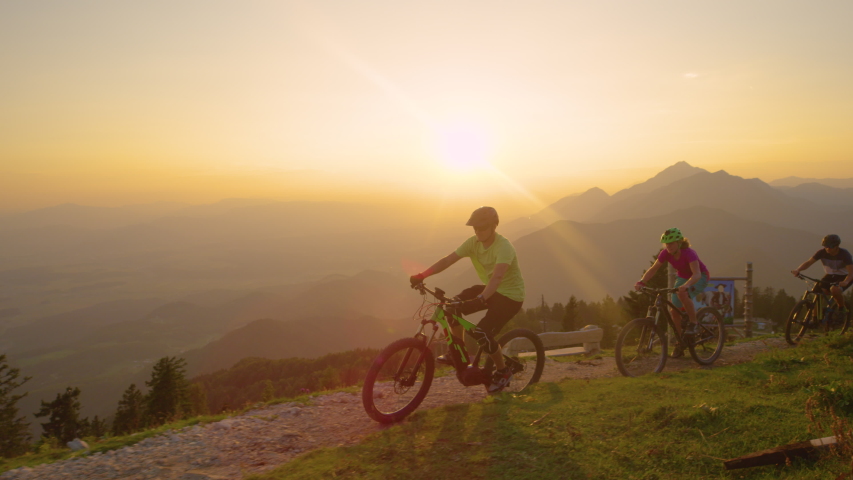 LENS FLARE, SLOW MOTION, DRONE: Cheerful active tourists riding their mountain bicycles up a scenic trail at sunset. Young travelers riding their electric bikes up a narrow mountain path at sunrise Royalty-Free Stock Footage #1029733649