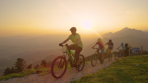 LENS FLARE, SLOW MOTION, DRONE: Cheerful active tourists riding their mountain bicycles up a scenic trail at sunset. Young travelers riding their electric bikes up a narrow mountain path at sunrise