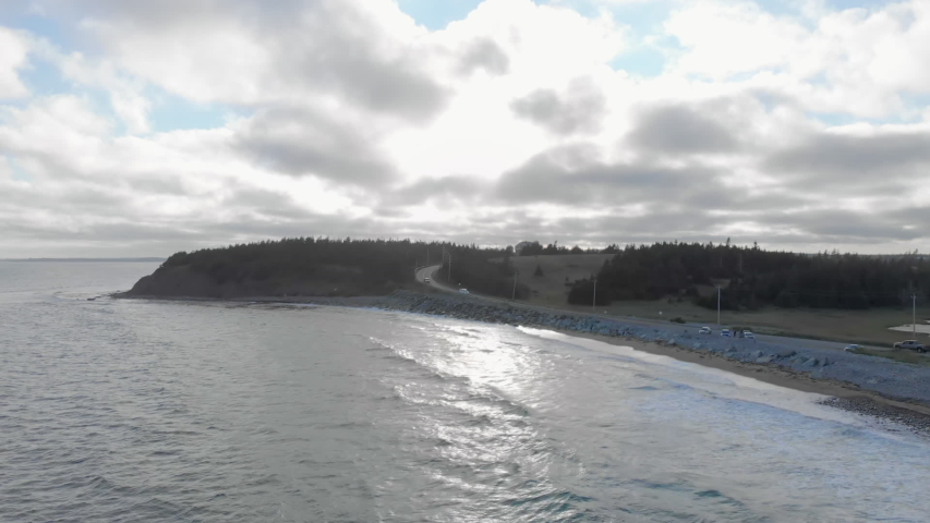 Cinematic drone - aerial footage moving forwards showing a road by the ocean and waves hitting the beach at Cole Harbour coast in Lawrencetown, Nova Scotia, Canada during summer season.