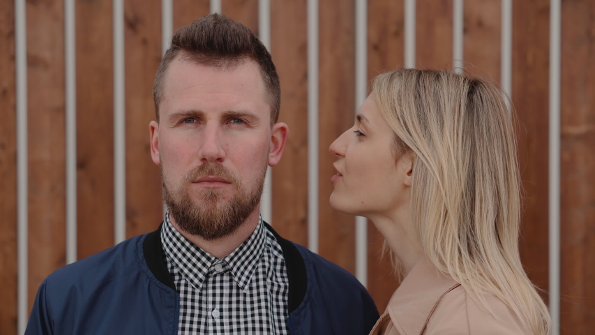 Charming lady is speaking silently in ear of young guy on street. He is looking forward calmly and listening her, medium shot
