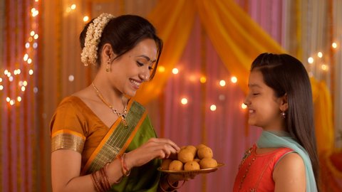 Smiling Indian mother feeding sweets to her young daughter during festivities - Happy Family. Indian stock video of a mother wishing daughter on the auspicious day of Diwali festival / Durga Puja