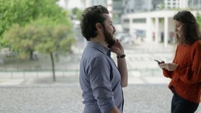 Side view of man and woman using cell phones outdoor. Smiling bearded man talking by smartphone while young woman passing him and texting with mobile phone. Technology concept